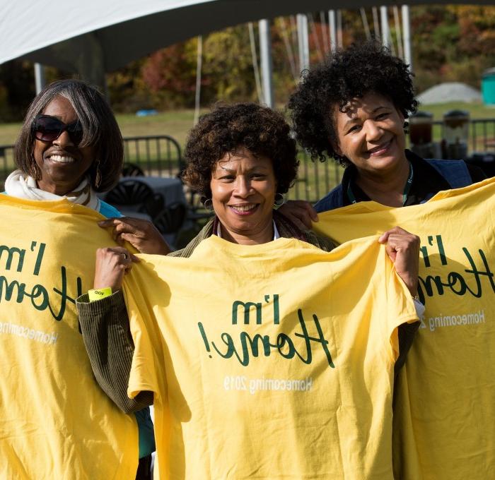 Three female alum at homecoming in end zone holding I'm 首页 t-shirts